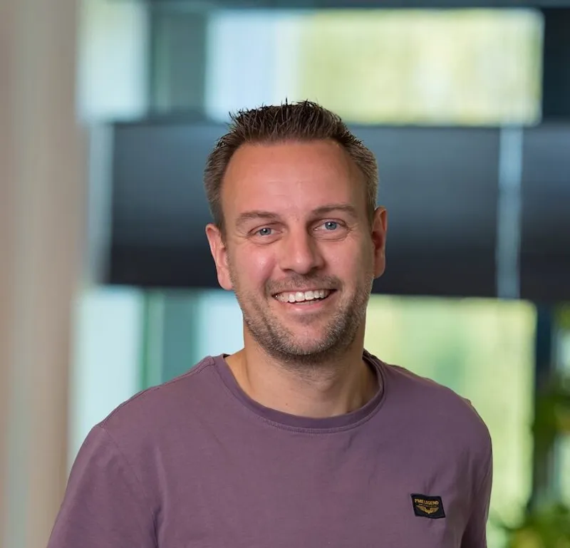 Portret van een man van begin veertig met kort blondbruin haar en stoppelbaard, gekleed in een paars T-shirt met logo, vriendelijk glimlachend op het Twyzer kantoor.