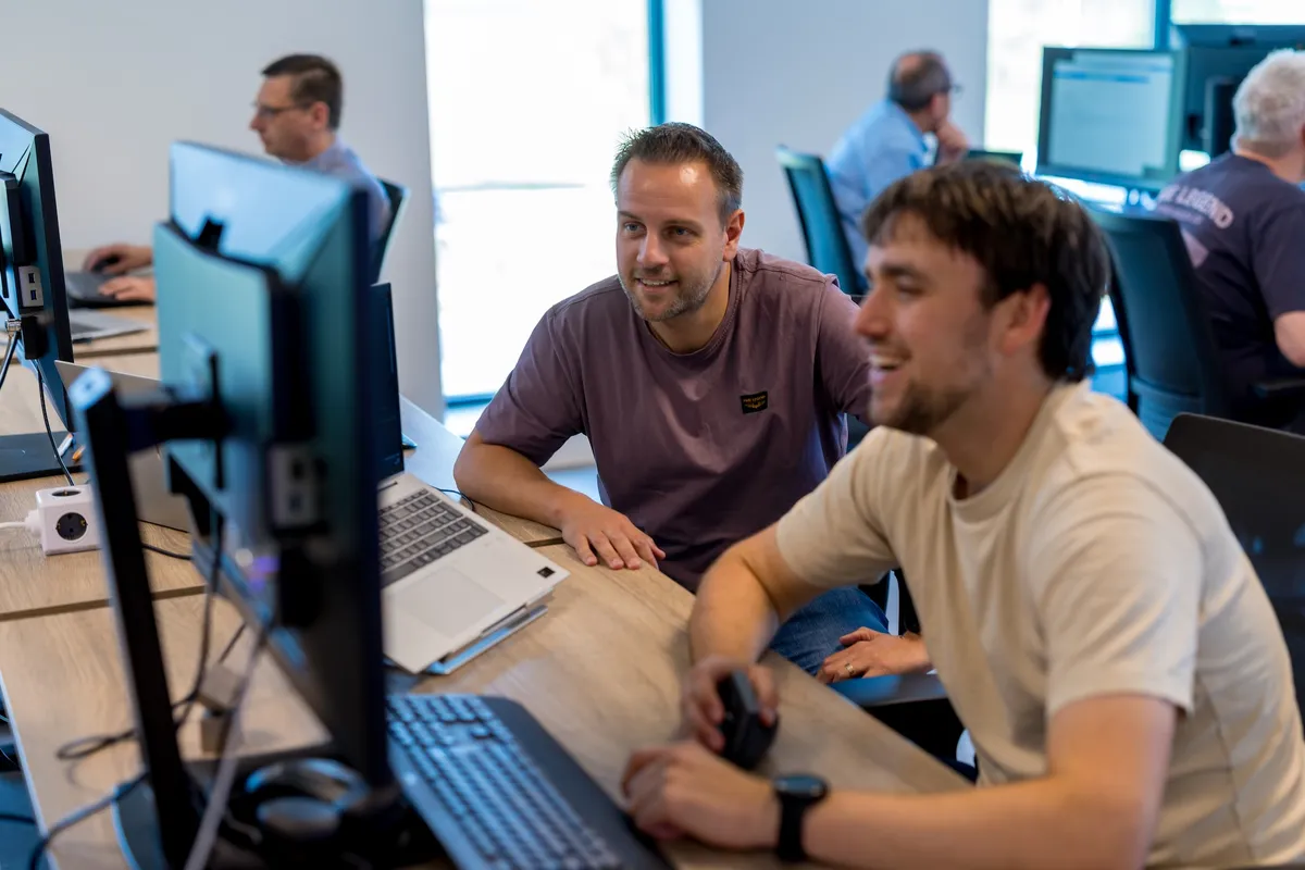 Collega’s samenwerken aan bureau Twee mannelijke medewerkers van Twyzer werken samen aan een computerscherm en lachen; een jongere man in een licht T-shirt en een oudere man in een paars T-shirt op een licht kantoor met collega's op de achtergrond.