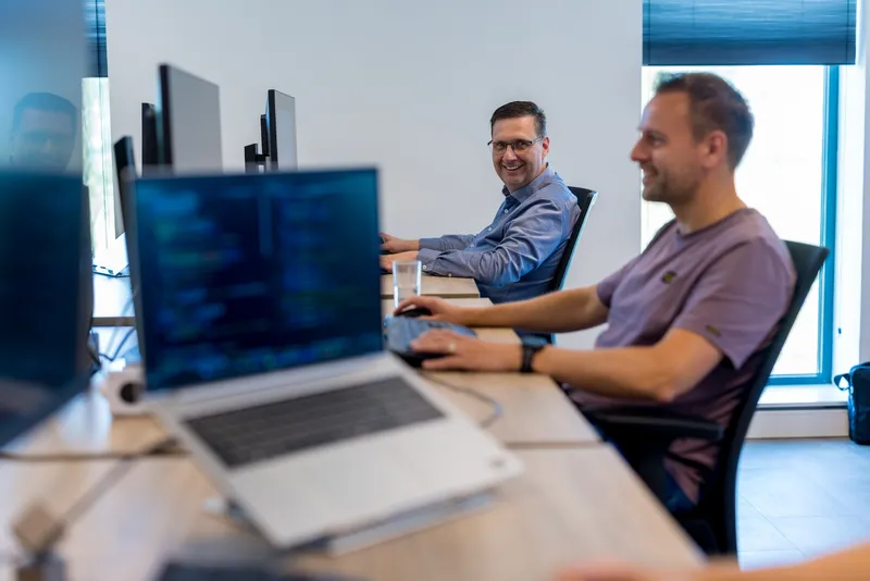 Op het Twyzer kantoor werkt een man in een paars T-shirt aan een laptop met code op de voorgrond, terwijl een man van middelbare leeftijd met bril en blauw overhemd glimlachend over zijn schouder kijkt.