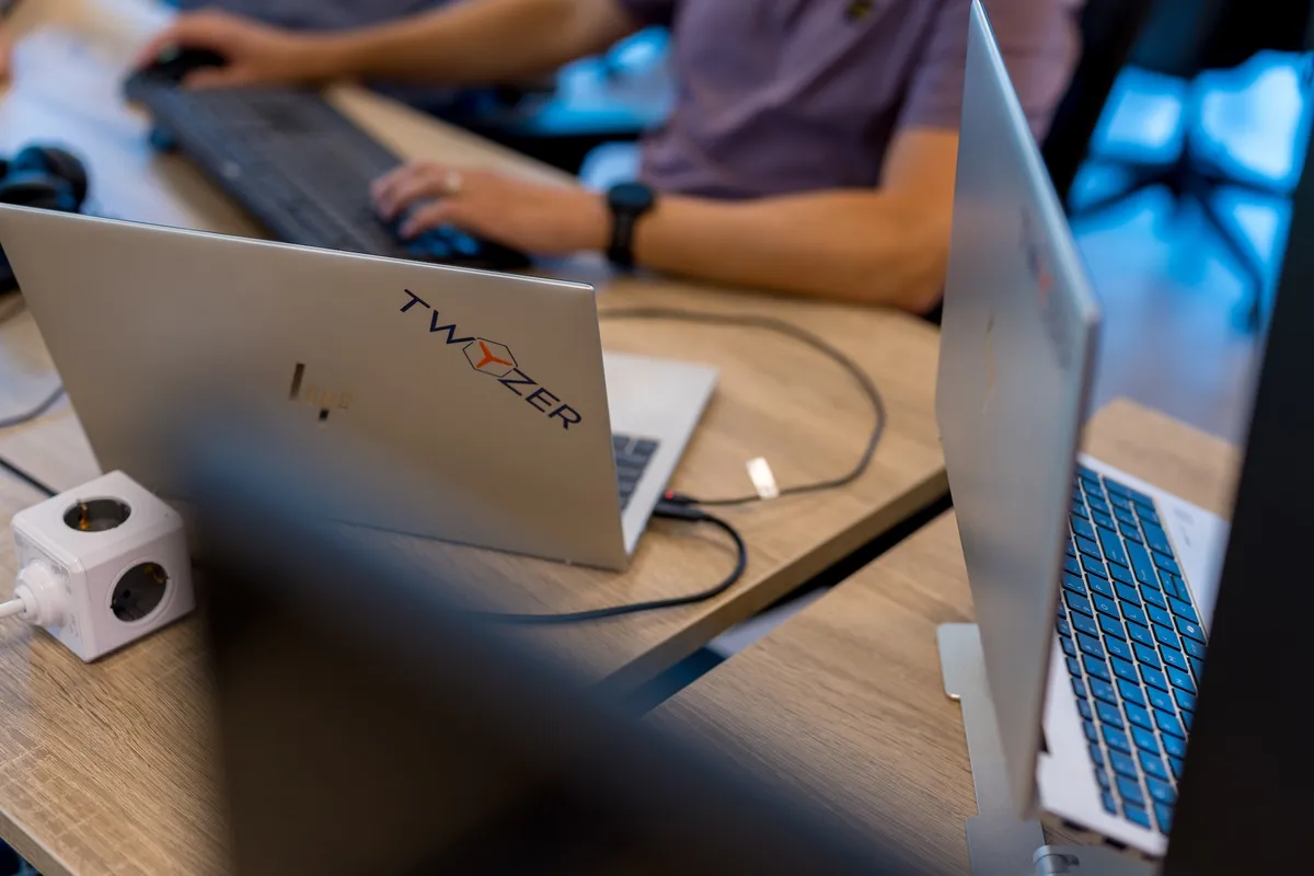 Laptop met Twyzer logo Close-up van een zilveren laptop met een Twyzer-sticker op een houten bureau, met op de achtergrond een medewerker in een paars poloshirt die aan een extern toetsenbord werkt.