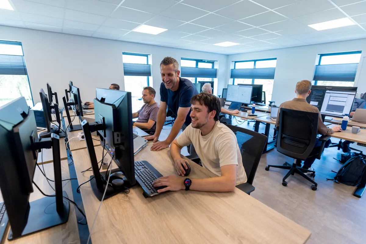 Open kantoor Twyzer samenwerking Brede opname van het open kantoor van Twyzer waar een jonge man in een crème T-shirt en een man van middelbare leeftijd in een blauw T-shirt samen aan een scherm werken, omringd door collega's aan hun werkplekken.