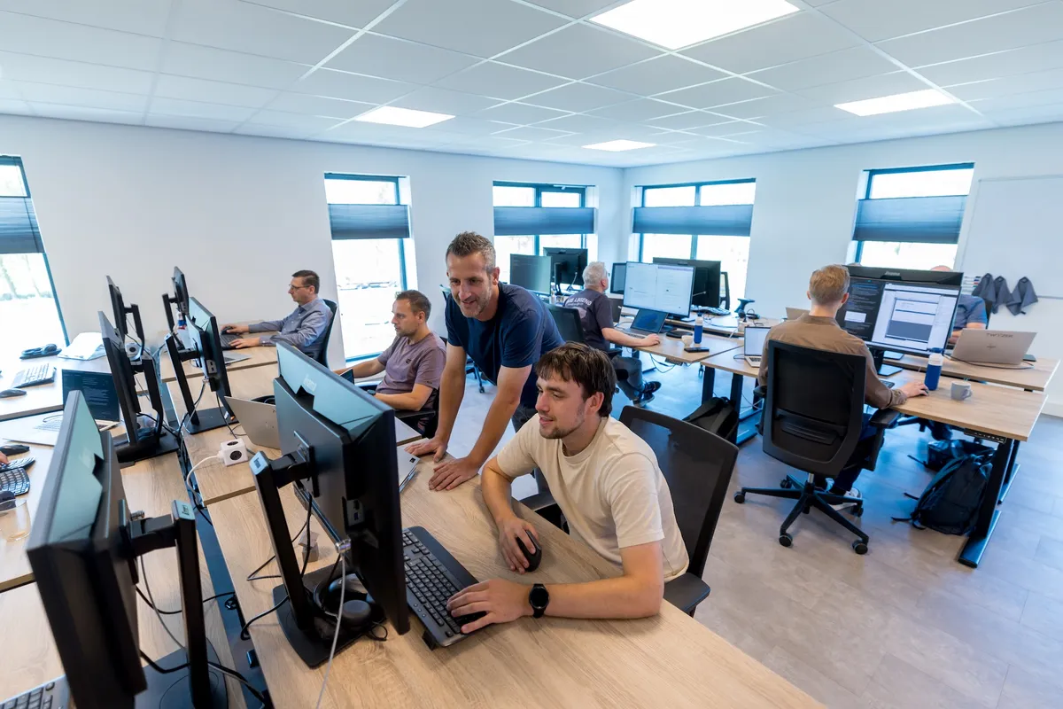 Kantoorruimte Twyzer team aan het werk Overzicht van het lichte open kantoor van Twyzer met meerdere medewerkers achter grote schermen; op de voorgrond werken een jonge man in een crème T-shirt en een man met grijs haar in een blauw T-shirt samen aan een monitor.