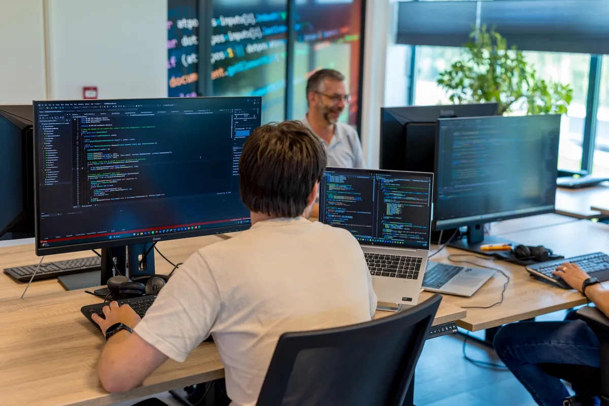 Softwareontwikkeling op kantoor Een jonge man in een beige T-shirt werkt aan programmeercode op meerdere schermen bij Twyzer, terwijl een oudere collega met grijs haar en bril op de achtergrond glimlacht in een licht modern kantoor.