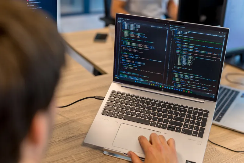 Close-up van een computerscherm met C#-code en Nederlandse commentaren in een donkere code-editor, gefotografeerd vanuit een hoek in een kantooromgeving.