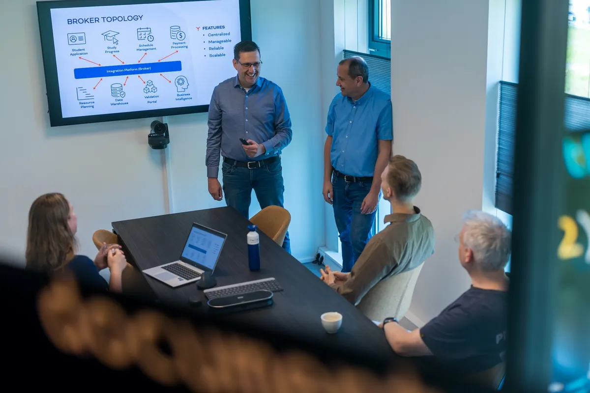 IT-presentatie achter glazen wand Een man presenteert een Broker Topology-diagram op een groot scherm aan zittende collega's rond een vergadertafel, gefotografeerd door een glazen scheidingswand.
