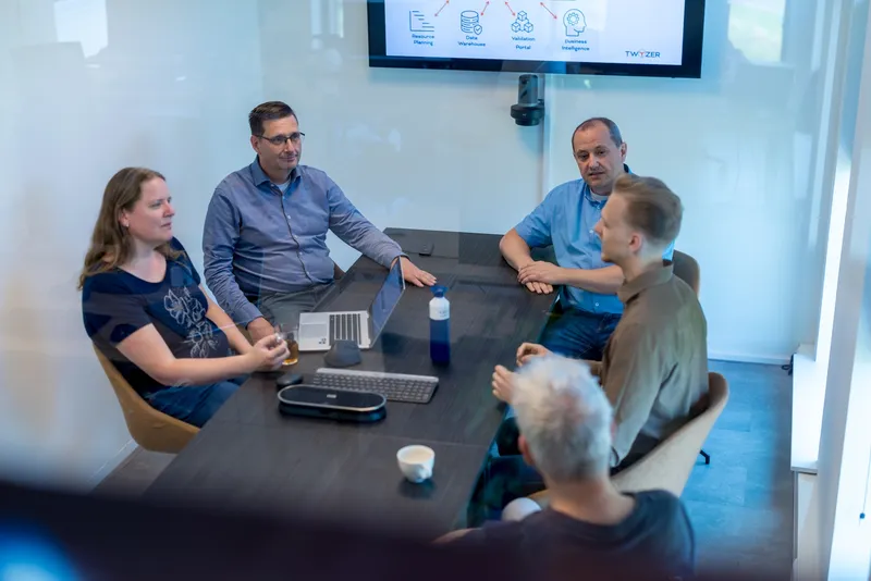 Vijf collega's zitten in een moderne vergaderzaal rond een tafel met laptop en conferentietelefoon, terwijl een technisch Twyzer-diagram op het scherm achter hen staat.