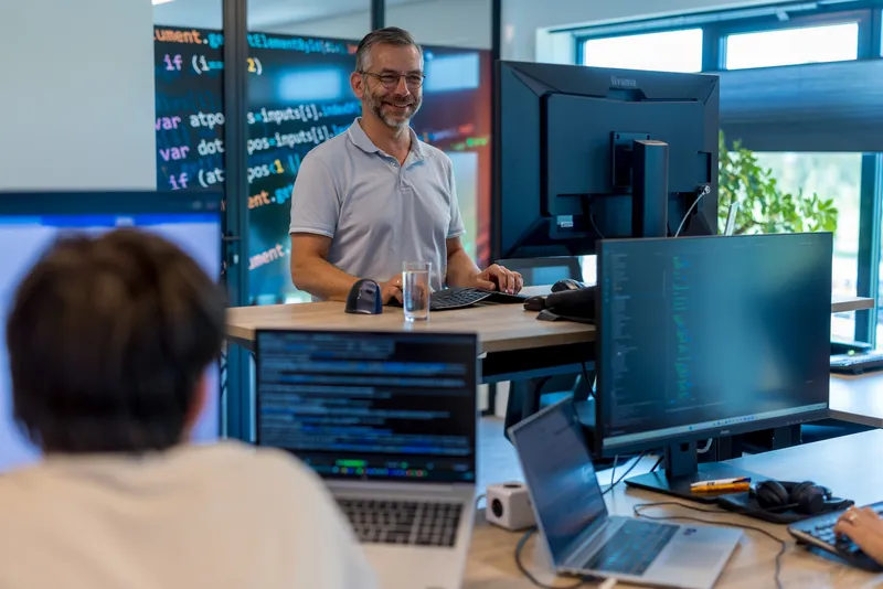Grip op Azure-kosten begint met inzicht en eigenaarschap Lachende collega achter een sta-bureau met meerdere schermen vol code