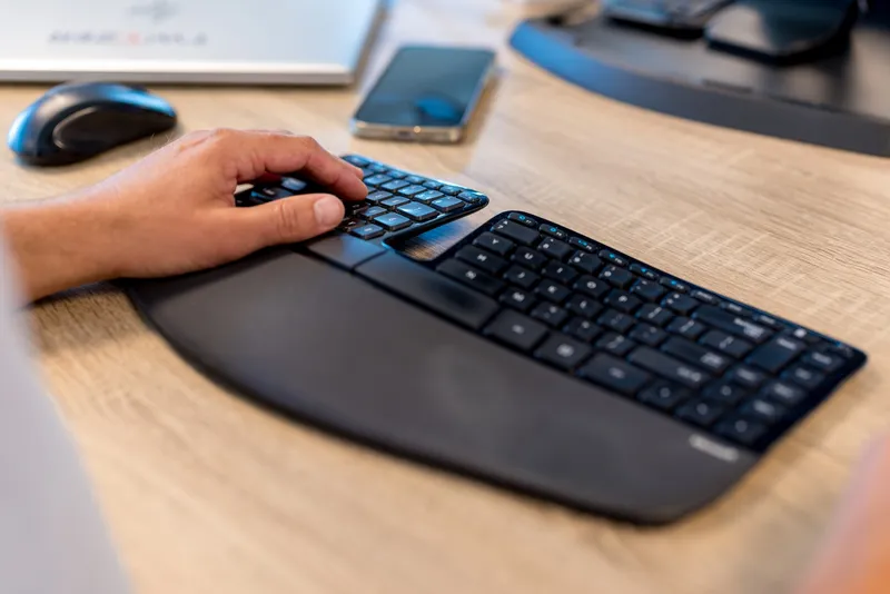 Close-up van een hand op een zwart ergonomisch split-toetsenbord op een houten bureau, met een muis, smartphone en monitor op de achtergrond.