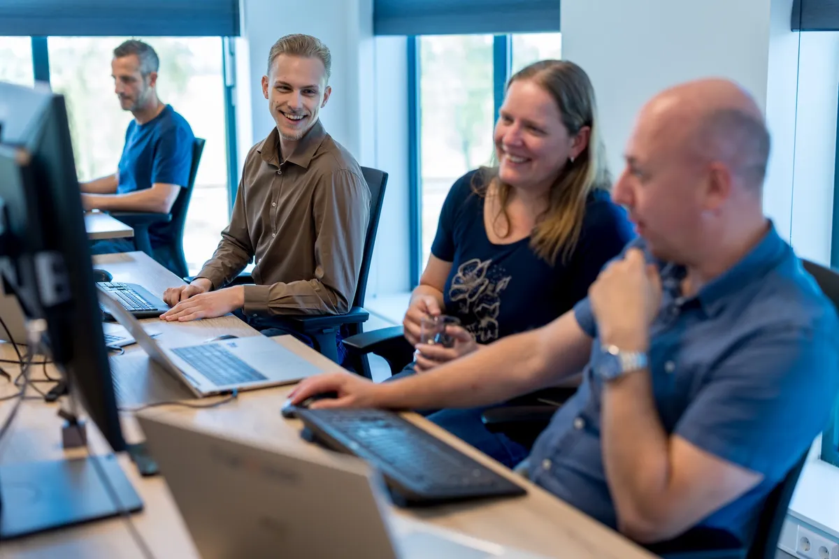Collega’s in gesprek op kantoor Vier medewerkers staan en zitten glimlachend bij hun bureaus in een licht kantoor en voeren een informeel gesprek met laptops en monitoren op de achtergrond.