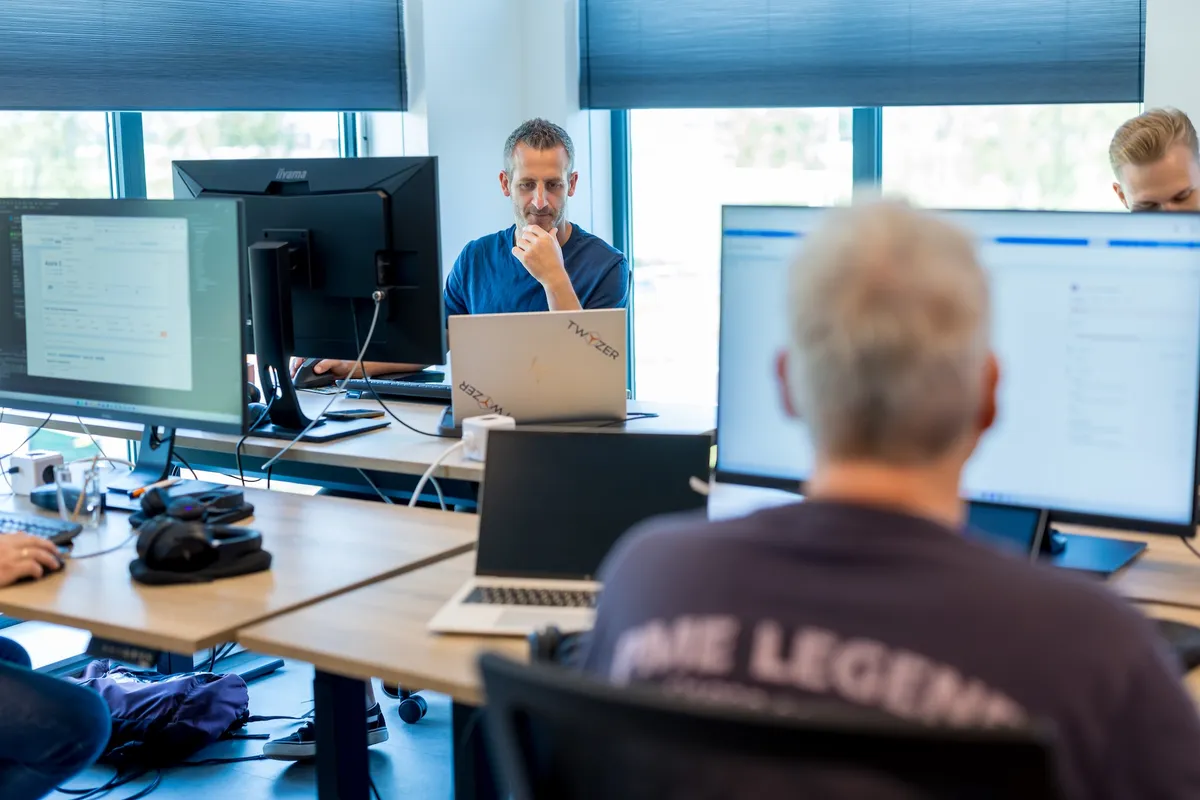 Medewerkers aan het werk op kantoor In een lichte kantoorruimte werken meerdere medewerkers geconcentreerd achter hun bureaus met laptops en monitoren; op de achtergrond een man met een Twyzer-laptop.