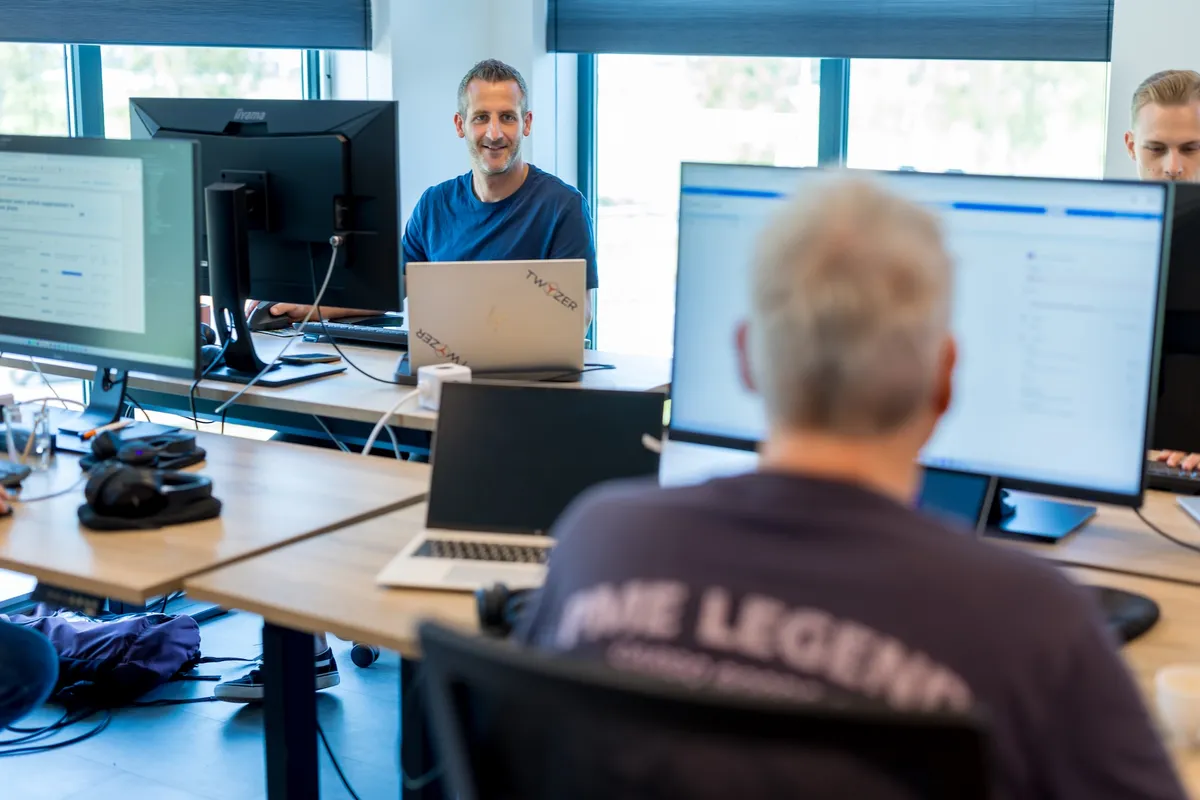 Glimlachende medewerker met Twyzer-laptop Een man in een blauw T-shirt zit glimlachend achter een laptop met Twyzer-stickers in een open kantoorruimte met grote ramen en blauwe jaloezieën.