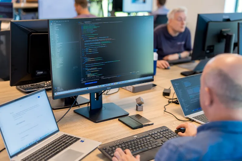 Een softwareontwikkelaar werkt aan een bureau met meerdere schermen waarop code en documentatie te zien zijn, terwijl collega's op de achtergrond aan het werk zijn.
